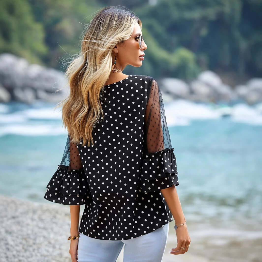 Woman wearing Black Polka Dot Sheer Blouse with 3/4 bell sleeves, standing by the beach, showcasing the blouse's elegant design.