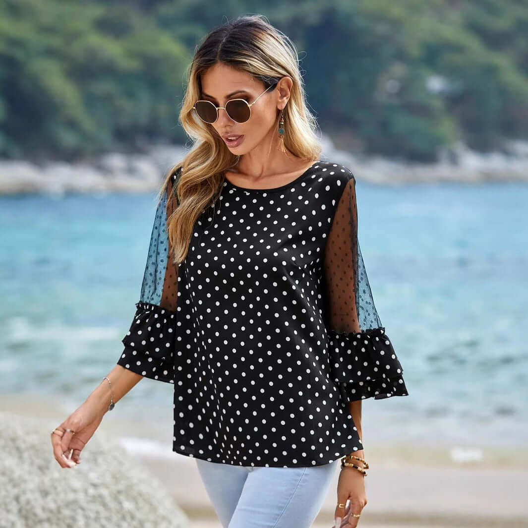 Woman wearing Black Polka Dot Sheer Blouse with 3/4 bell sleeves, styled with sunglasses by the sea. Fashionable and elegant look.