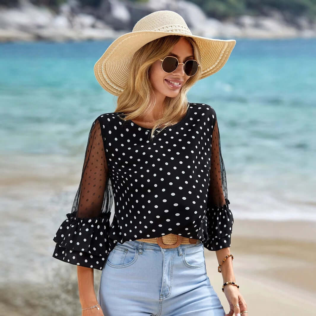 Woman wearing a Black Polka Dot Sheer Blouse with 3/4 bell sleeves, styled with jeans and sunglasses at the beach, exuding elegance.